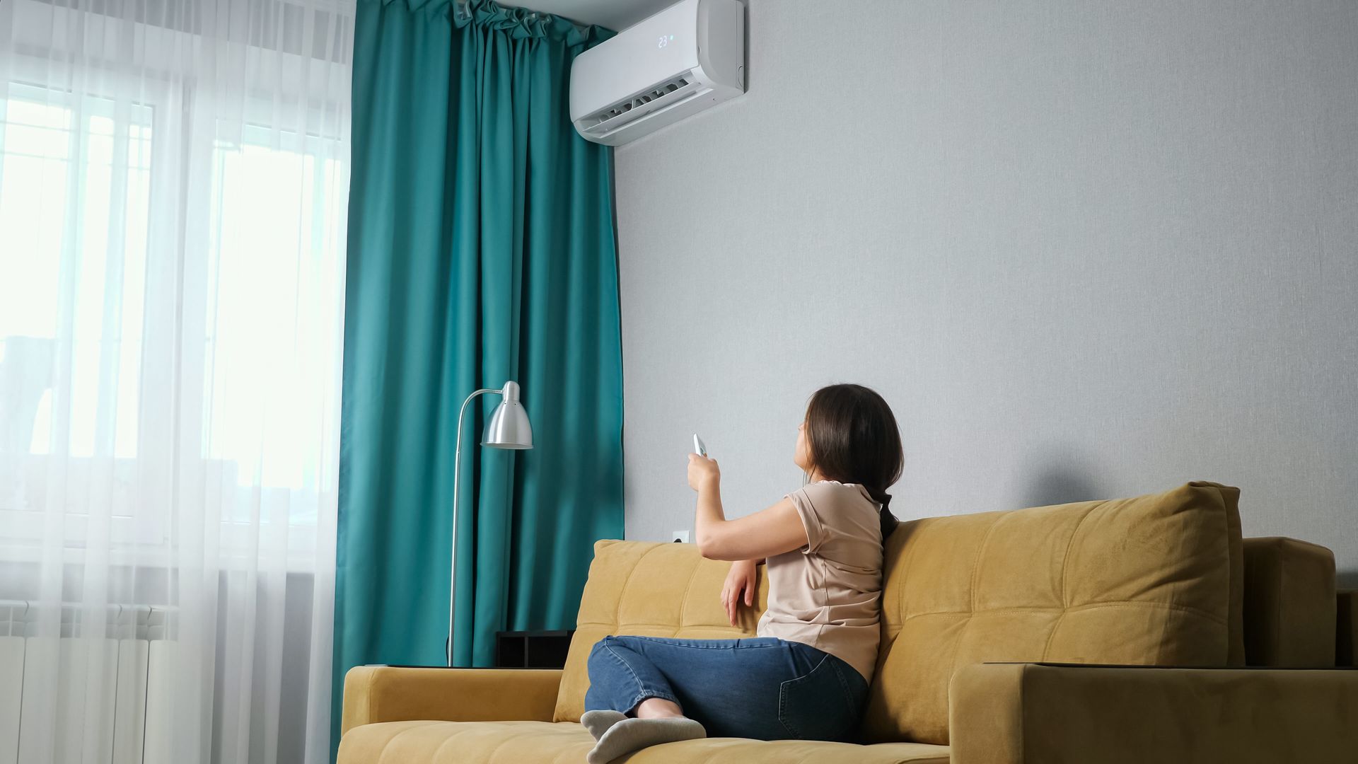 Woman sitting on a yellow sofa, holding a smartphone in a bright living room with teal curtains and an air conditioner.
