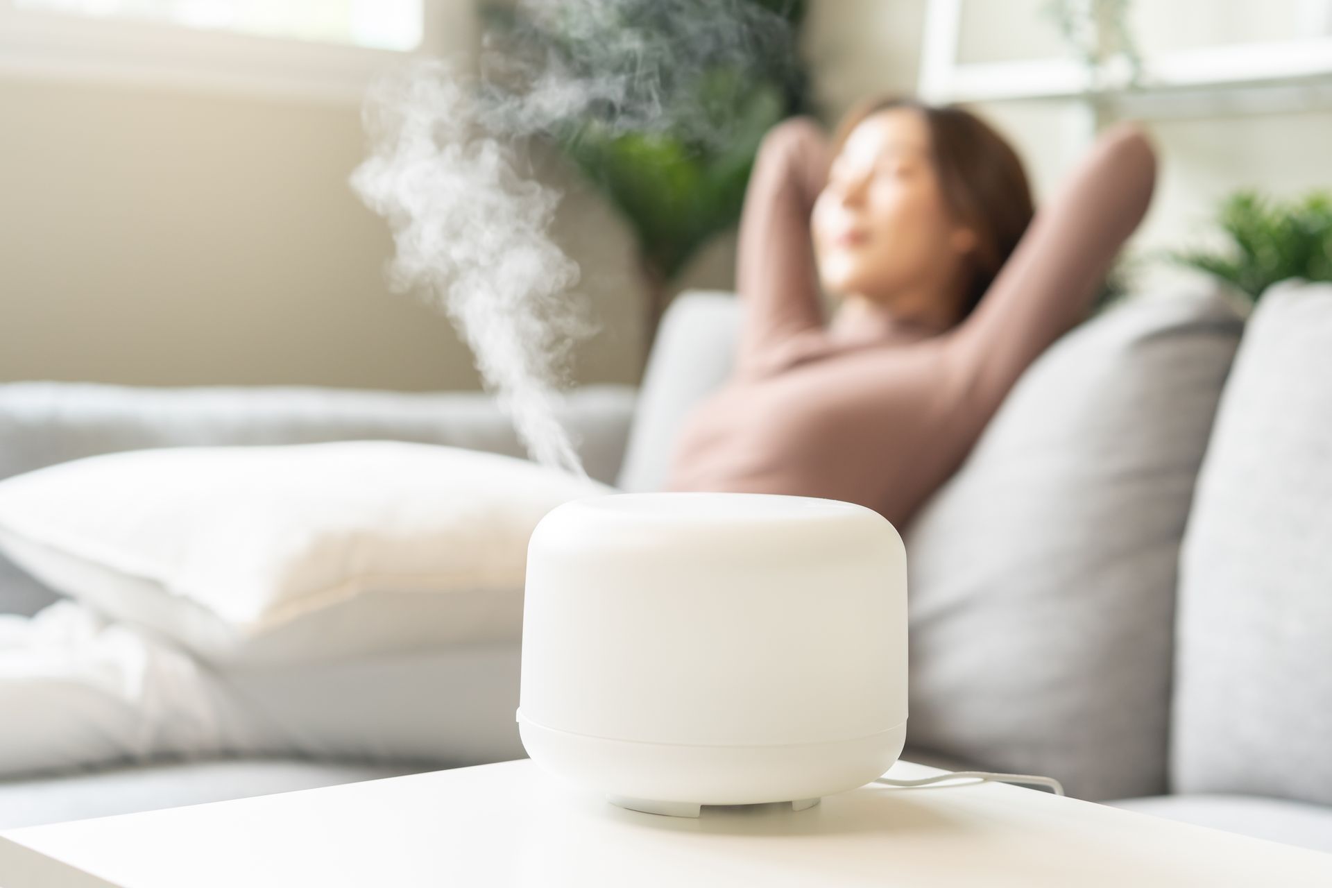 White essential oil diffuser steaming, woman relaxed on sofa in background.