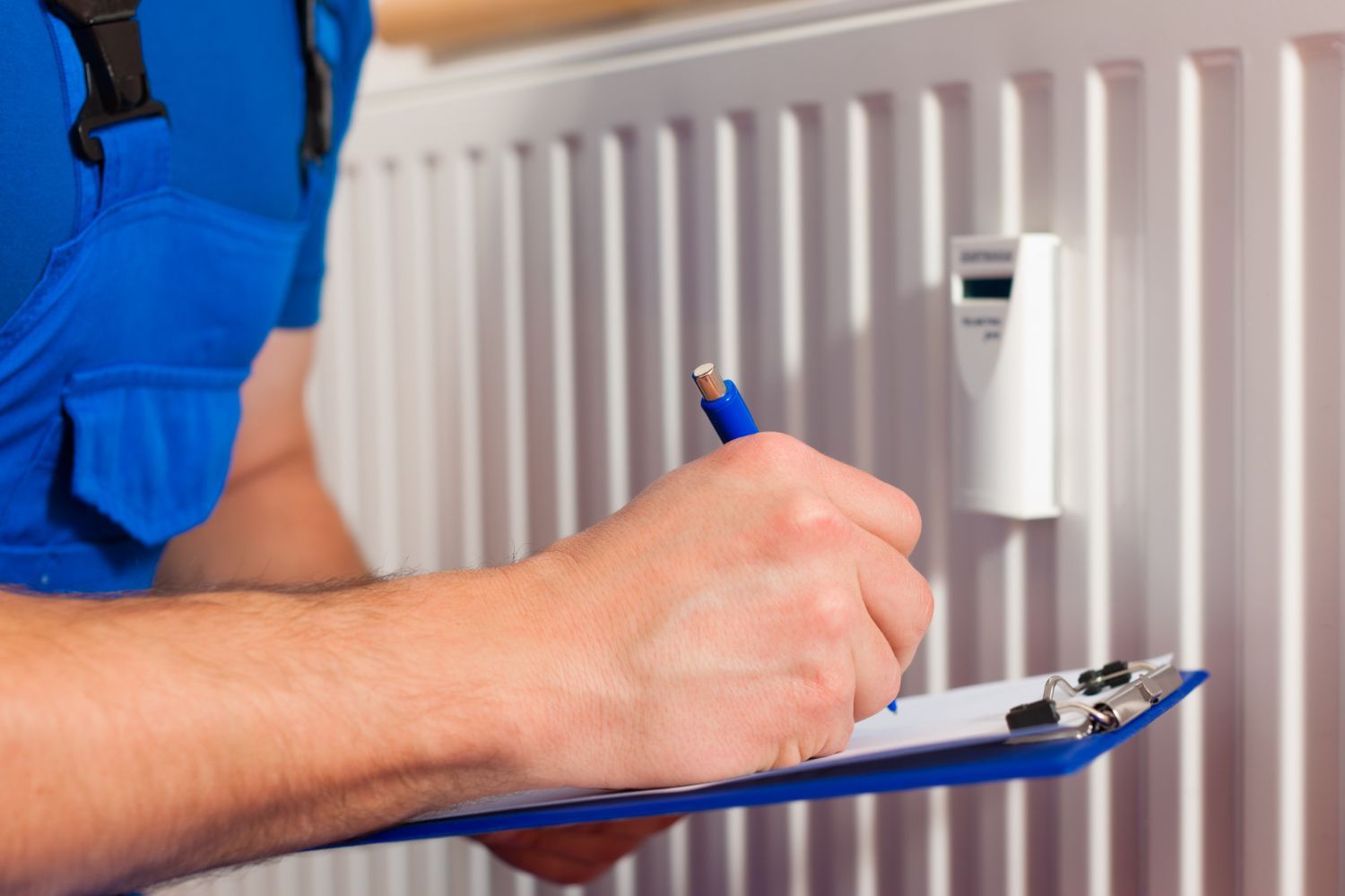 Person in blue overalls taking notes on a clipboard in front of a white radiator.