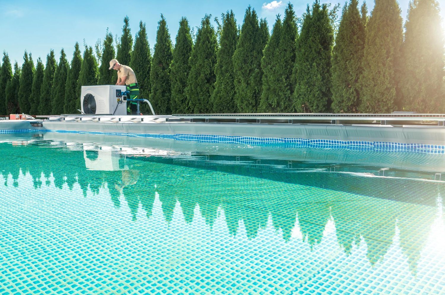 Man working on pool equipment beside a swimming pool, sunny day.