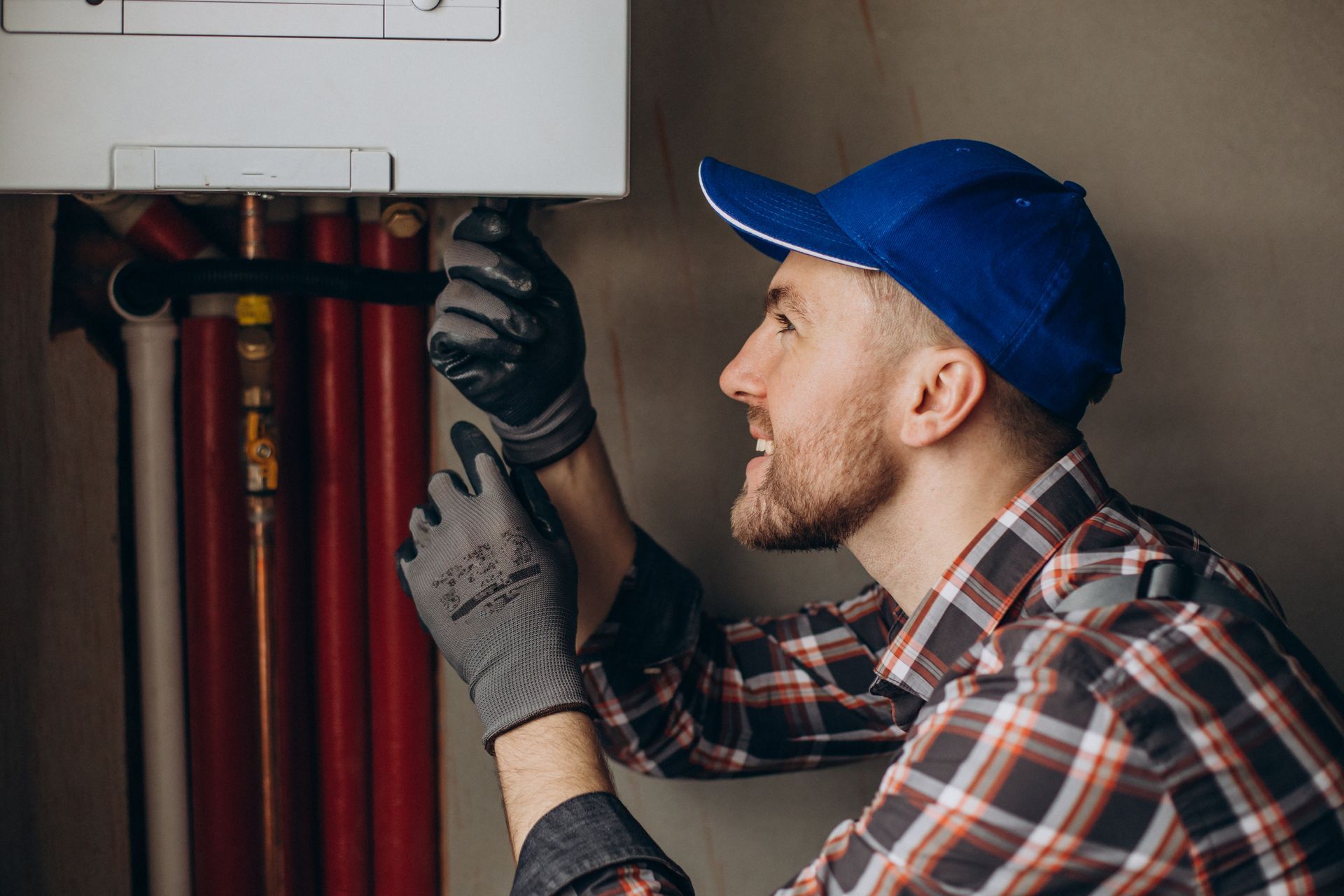 a man in a plaid shirt and blue hat is fixing a boiler .