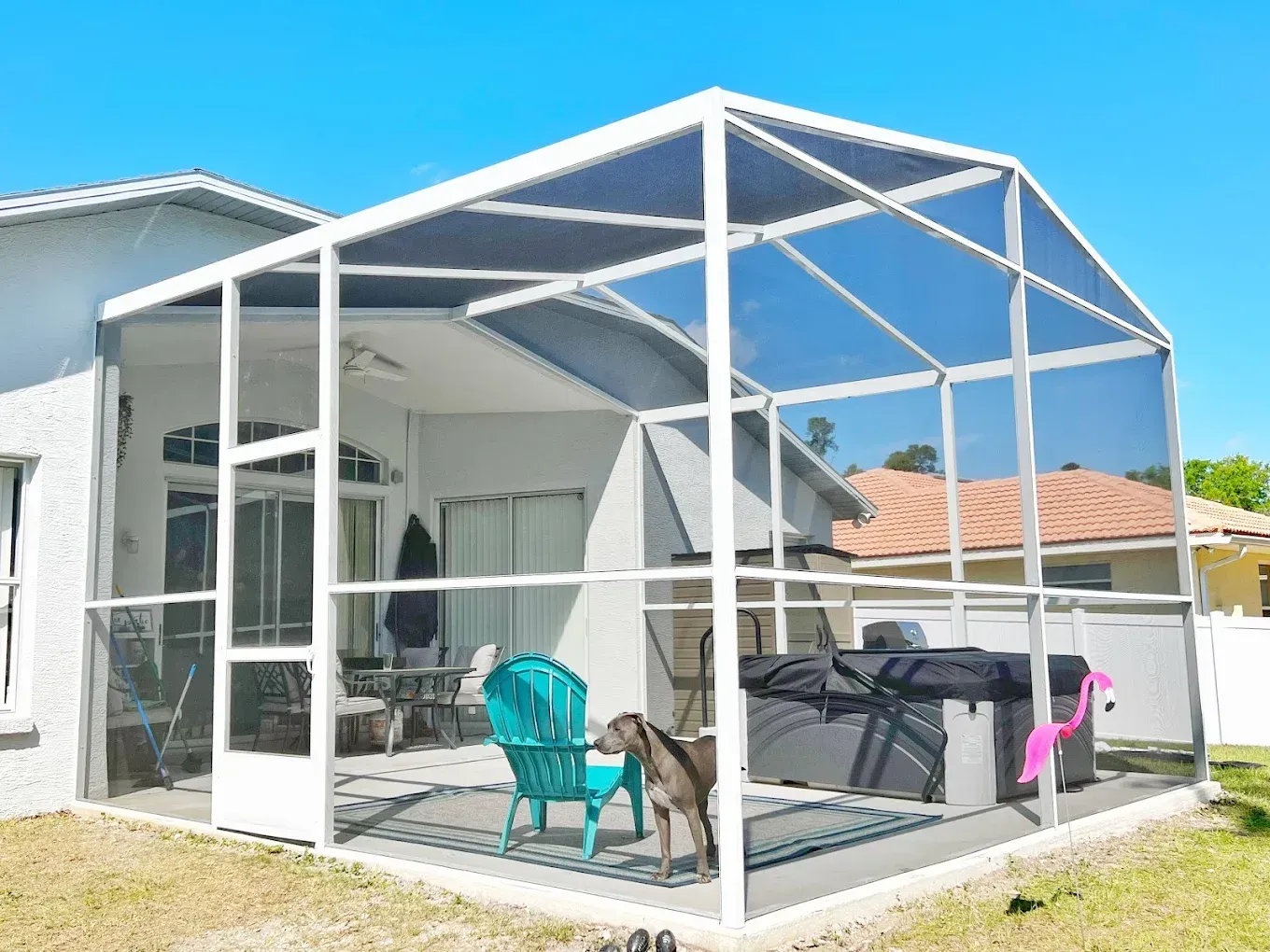 Screened-in patio with a dog, teal chair, grill, and house in the background under a blue sky.