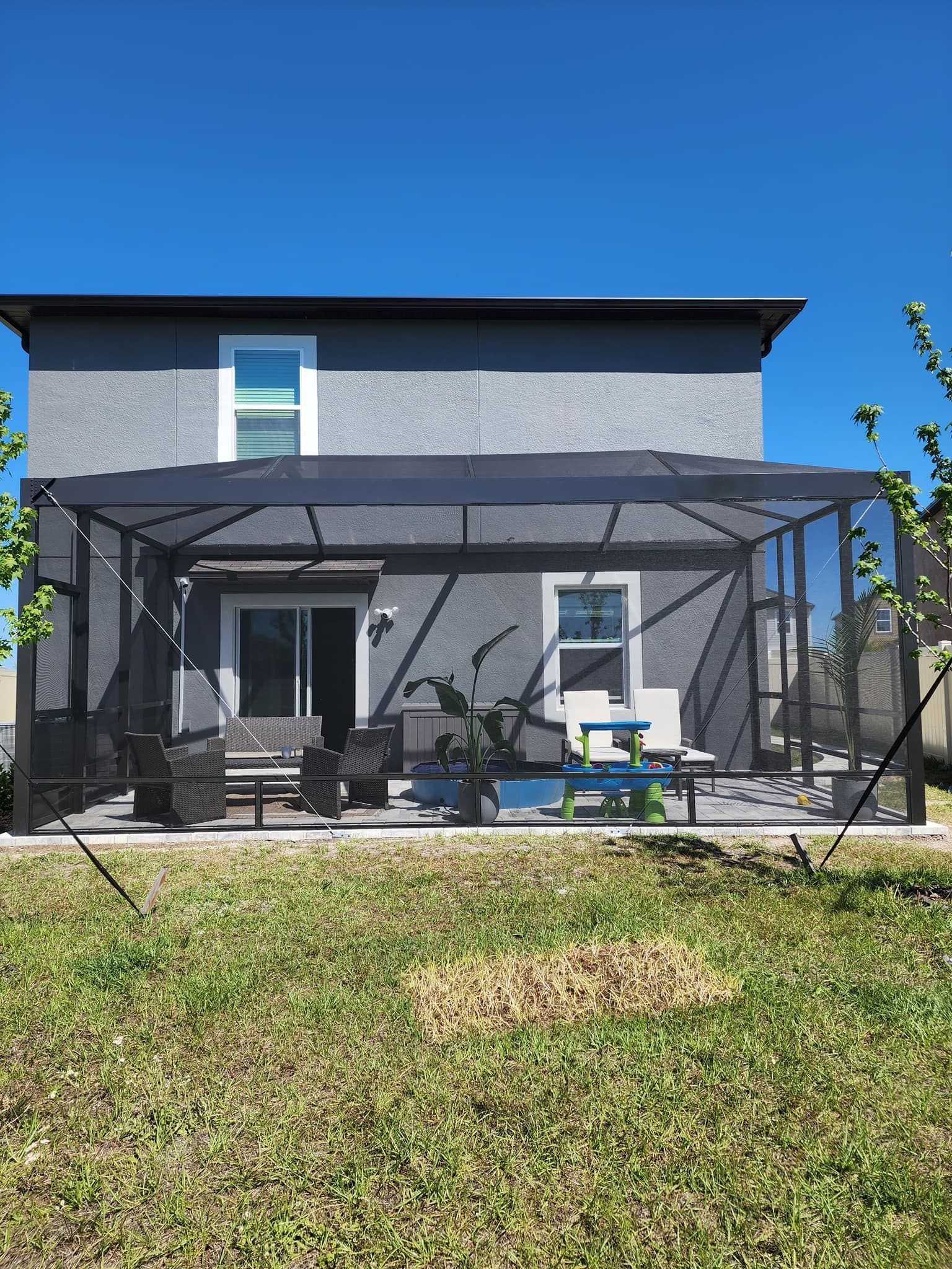 Back of a gray house with a black screened patio, chairs, and plants on a concrete patio, with blue sky.