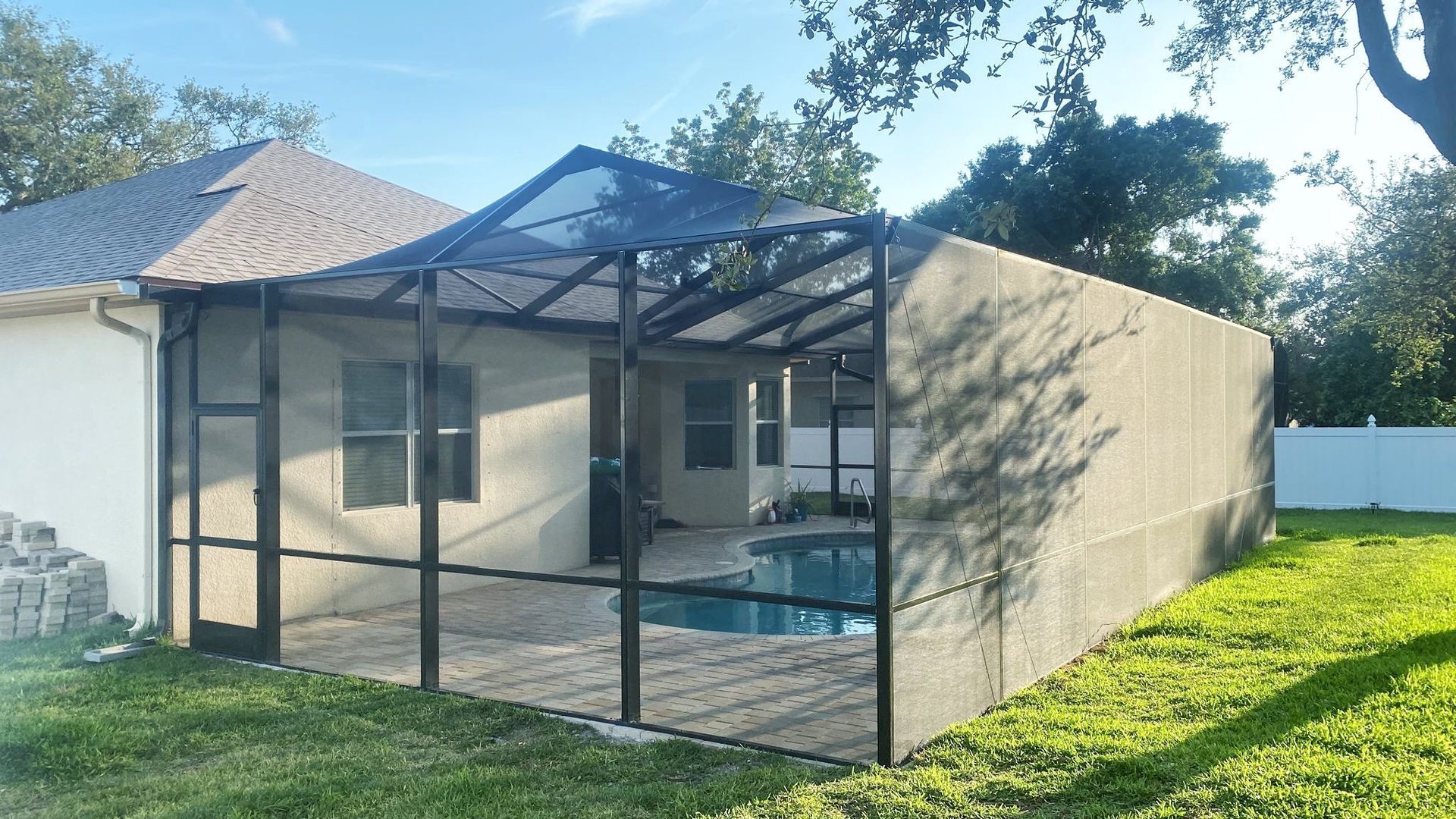 Pool enclosure attached to a house, with a pool visible inside. Green grass and trees are in the background.