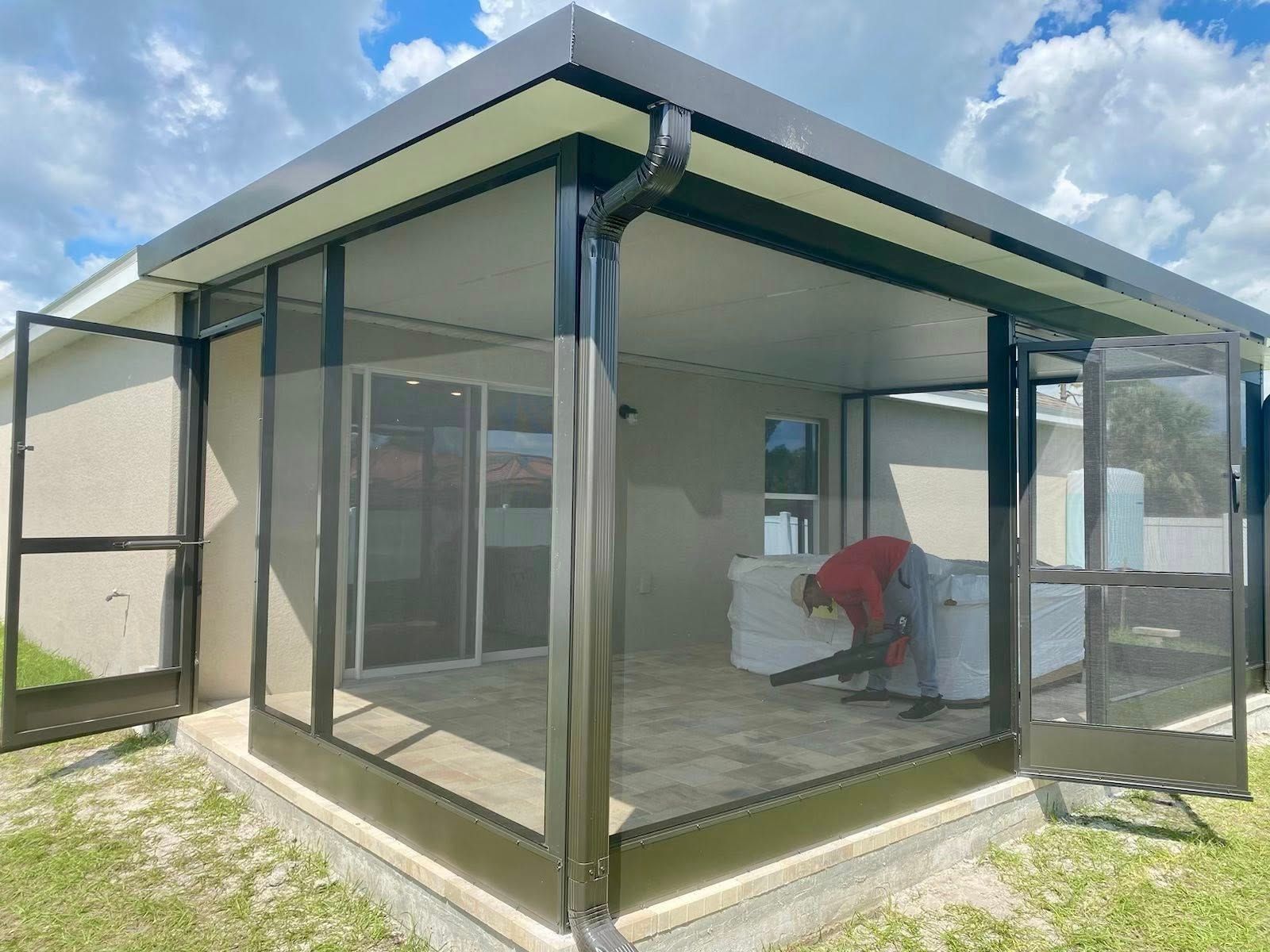 A screened-in patio with black framing, open doors, and a person using a leaf blower.