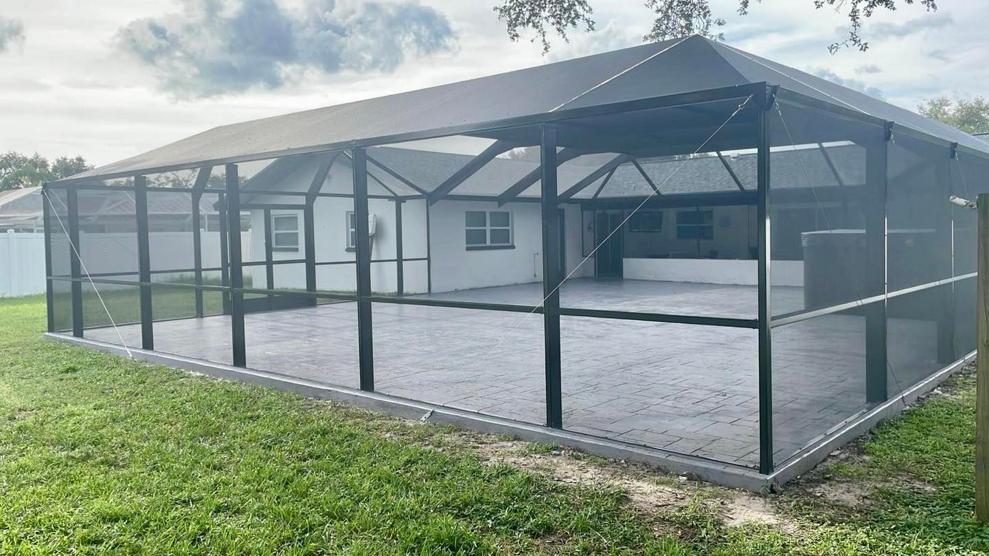 Screened-in patio enclosure with a gray concrete floor, attached to a white house, in a grassy backyard.