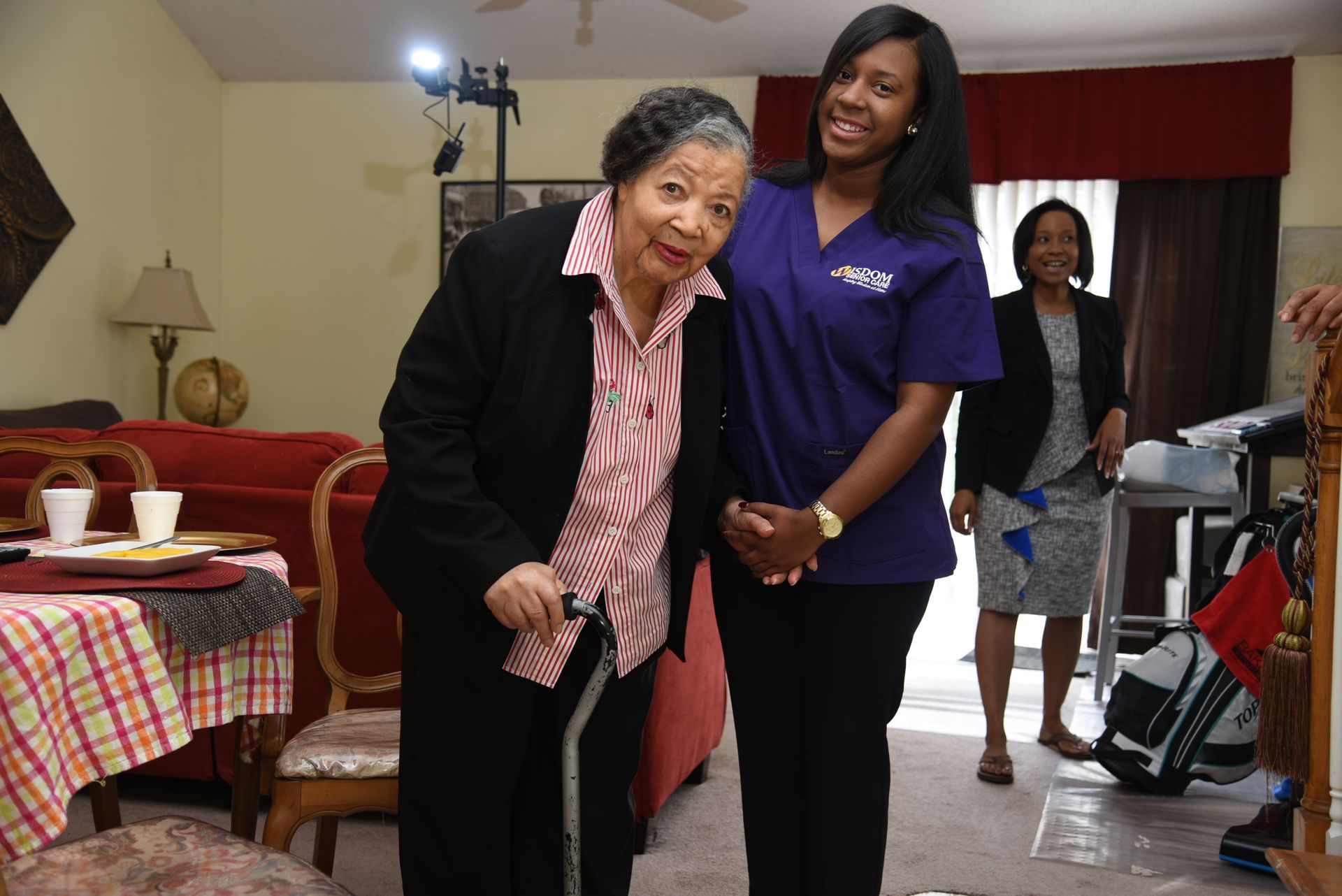 An elderly person with a cane smiles, posed with a woman in a purple uniform. A third woman stands in the background.