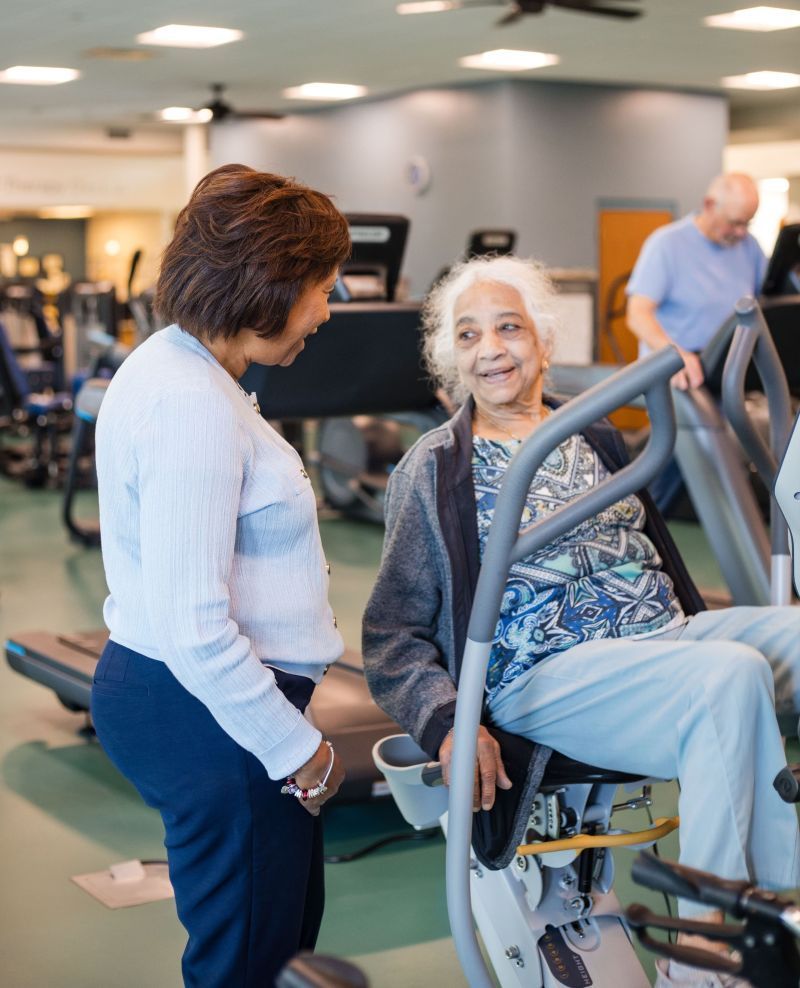 Woman assisting an older person on a recumbent exercise bike in a gym setting.