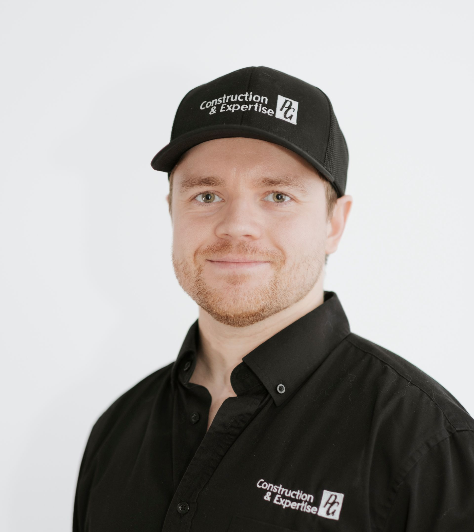 Homme en chemise et casquette noires sur fond blanc, esquissant un léger sourire