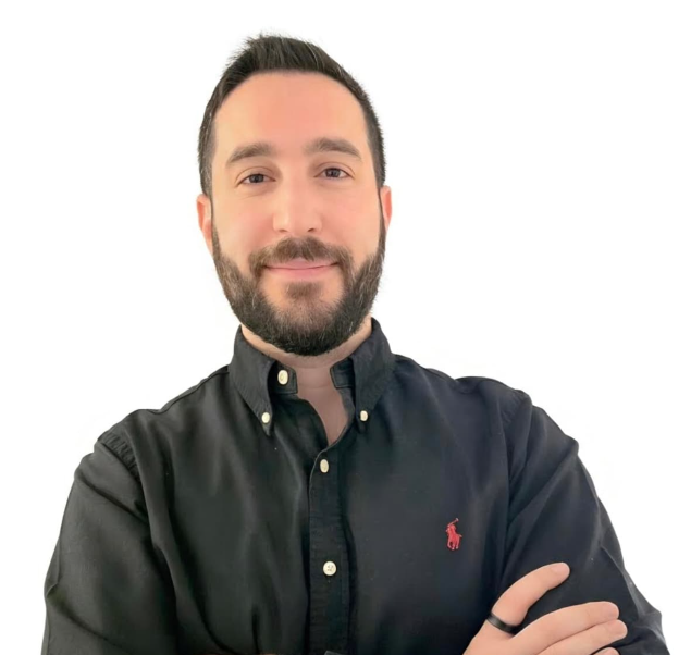Homme en chemise noire boutonnée, les bras croisés, souriant sur fond blanc