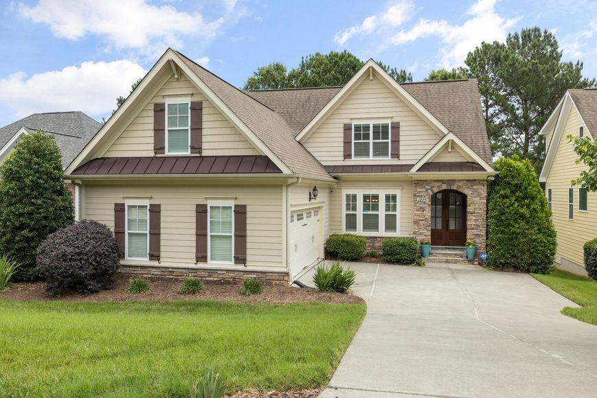 Tan house with brown trim, shutters, and a stone entryway, driveway, and green lawn.