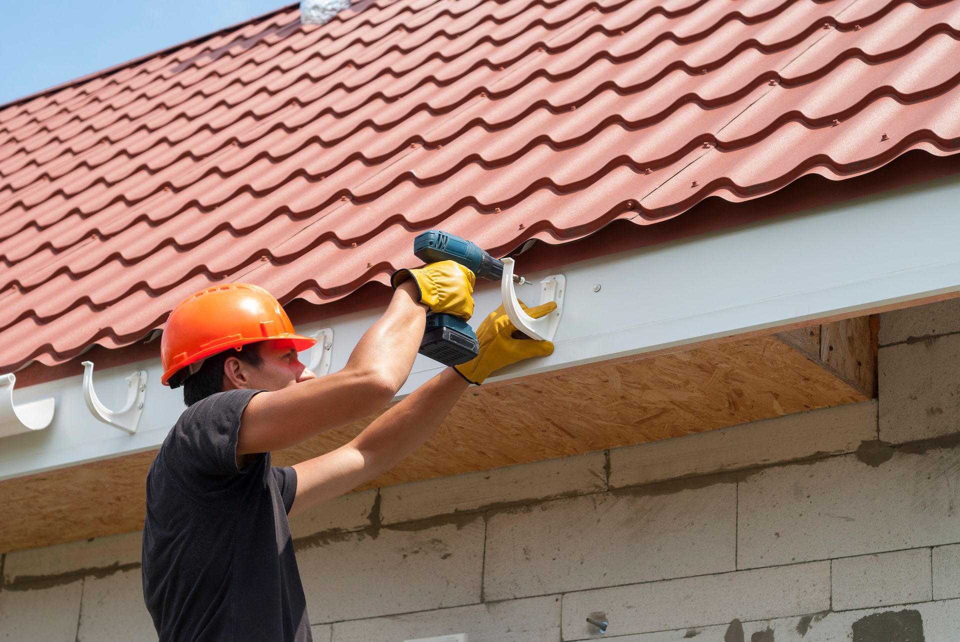 Worker installing white rain gutter on metal tile roof using drill during residential construction.