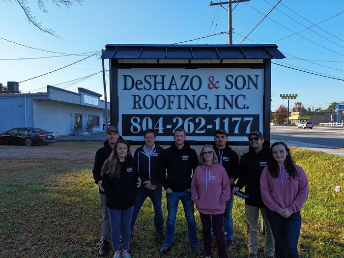 A group of people standing in front of a sign for deshazo & son roofing inc. A group of people standing in front of a sign for deshazo & son roofing inc.