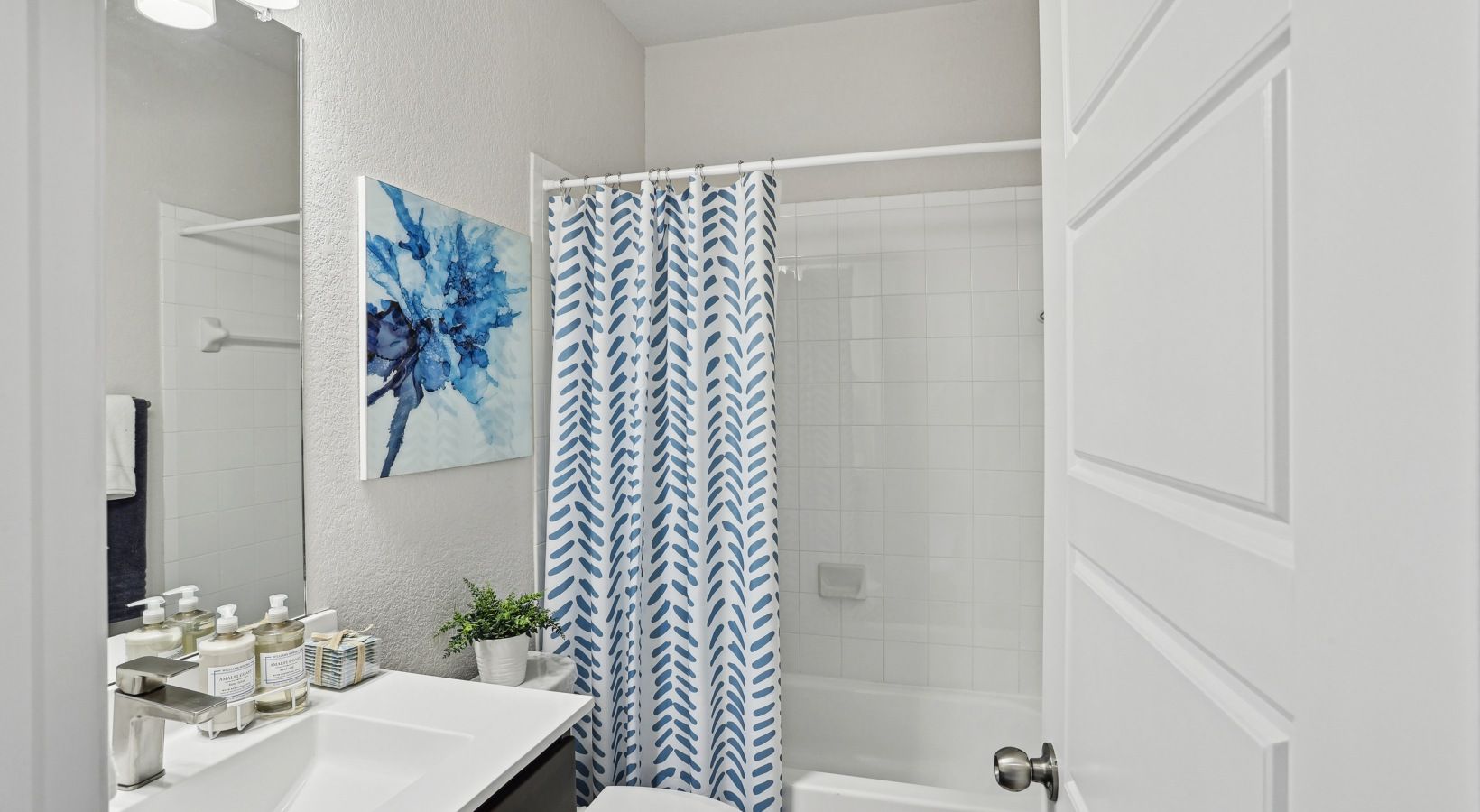 Bathroom with white tile shower, blue leaf-pattern curtain, and a sink with toiletries.