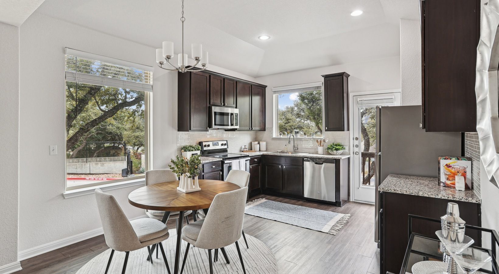 Modern apartment kitchen with dark wood cabinets, stainless appliances, and a round dining table by large windows.