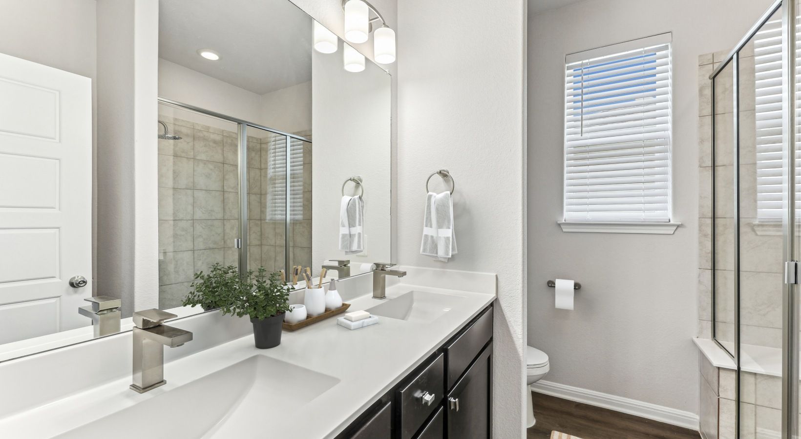 Bathroom with double vanity, glass shower enclosure, toilet, and window with blinds.