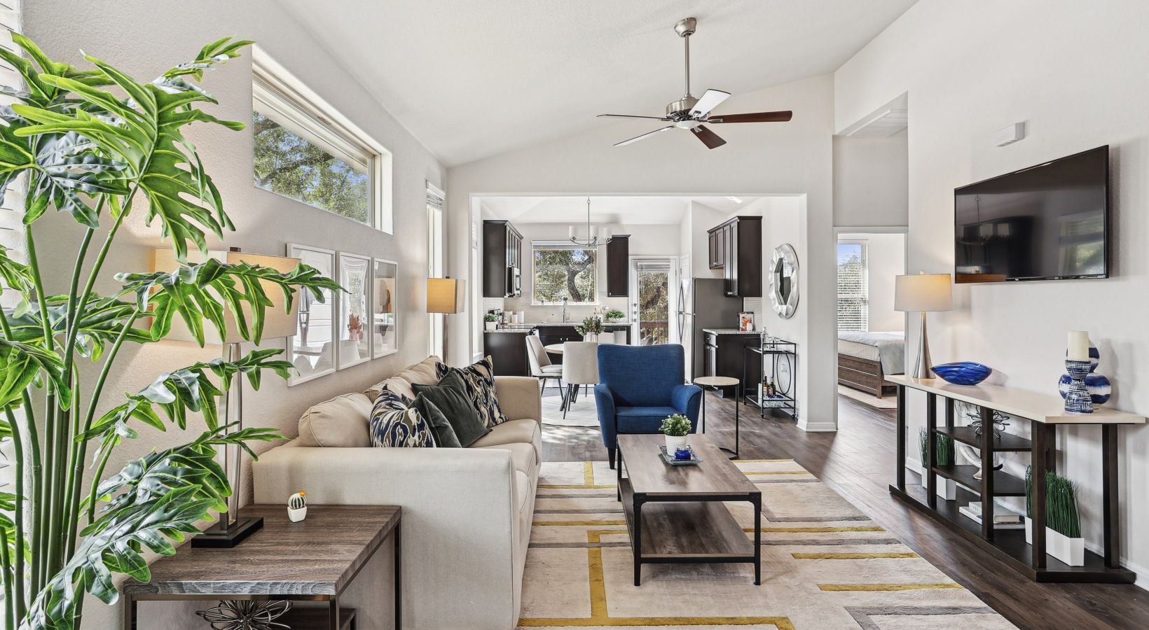 Open-concept living room with beige sofa, blue armchair, wall-mounted TV, ceiling fan, and kitchen in background.
