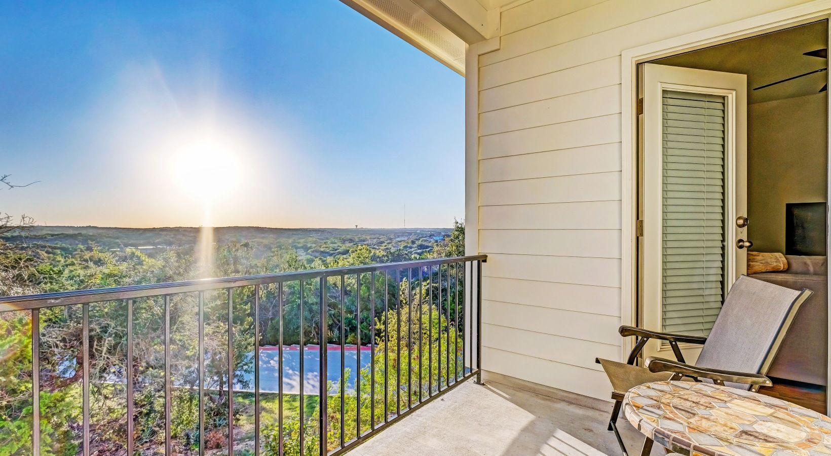 Outdoor balcony with railing, a chair and small mosaic table, overlooking trees and distant hills.