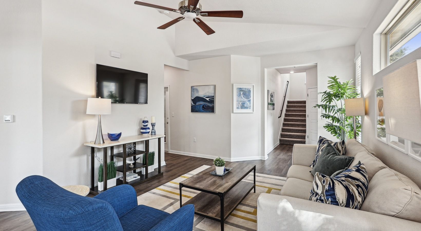 Living room in an apartment with a beige sofa, blue chair, coffee table, wall-mounted TV, ceiling fan, and staircase.