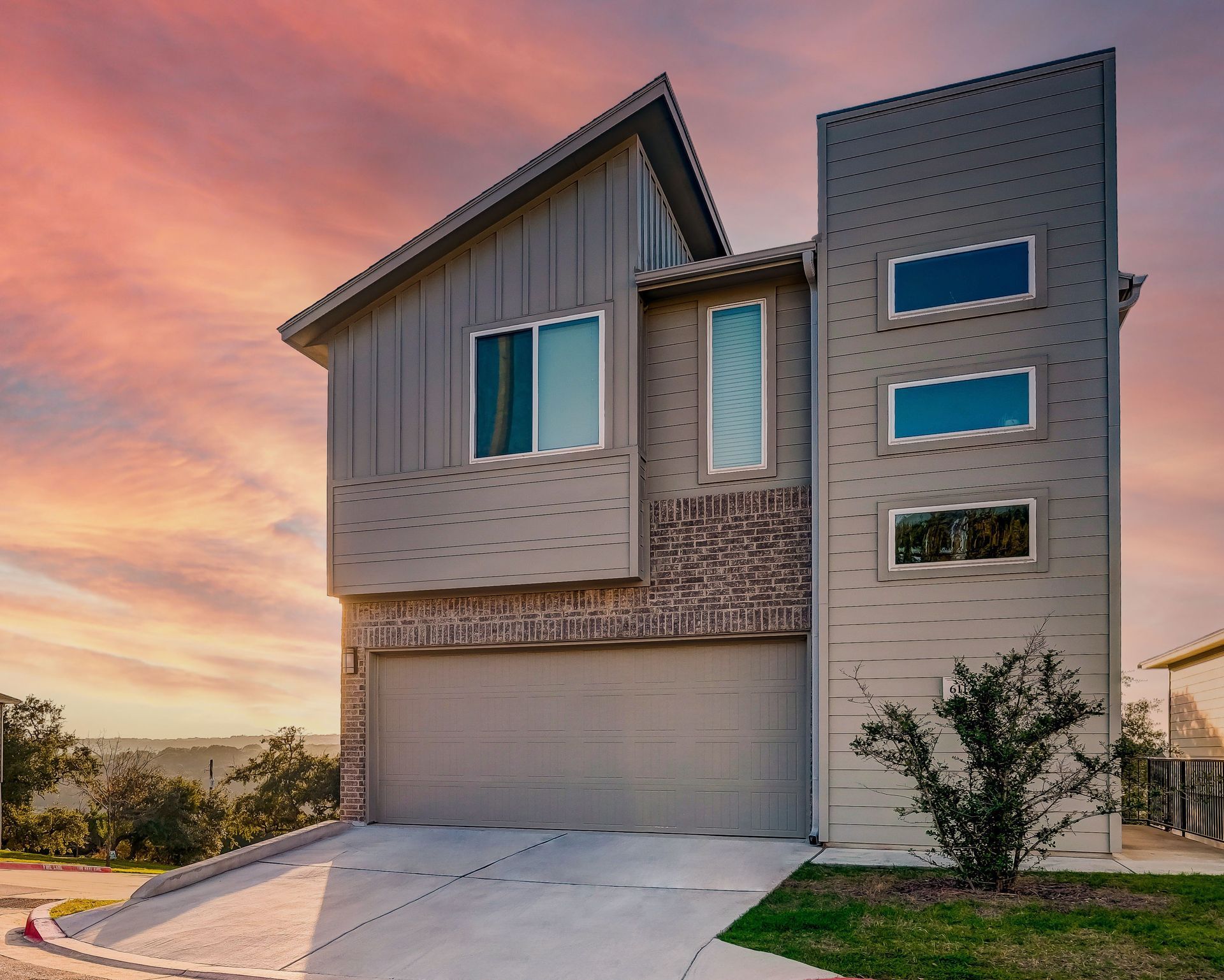 Modern two-story house with gray siding, brick accents, and a garage. Set against a sunset sky.