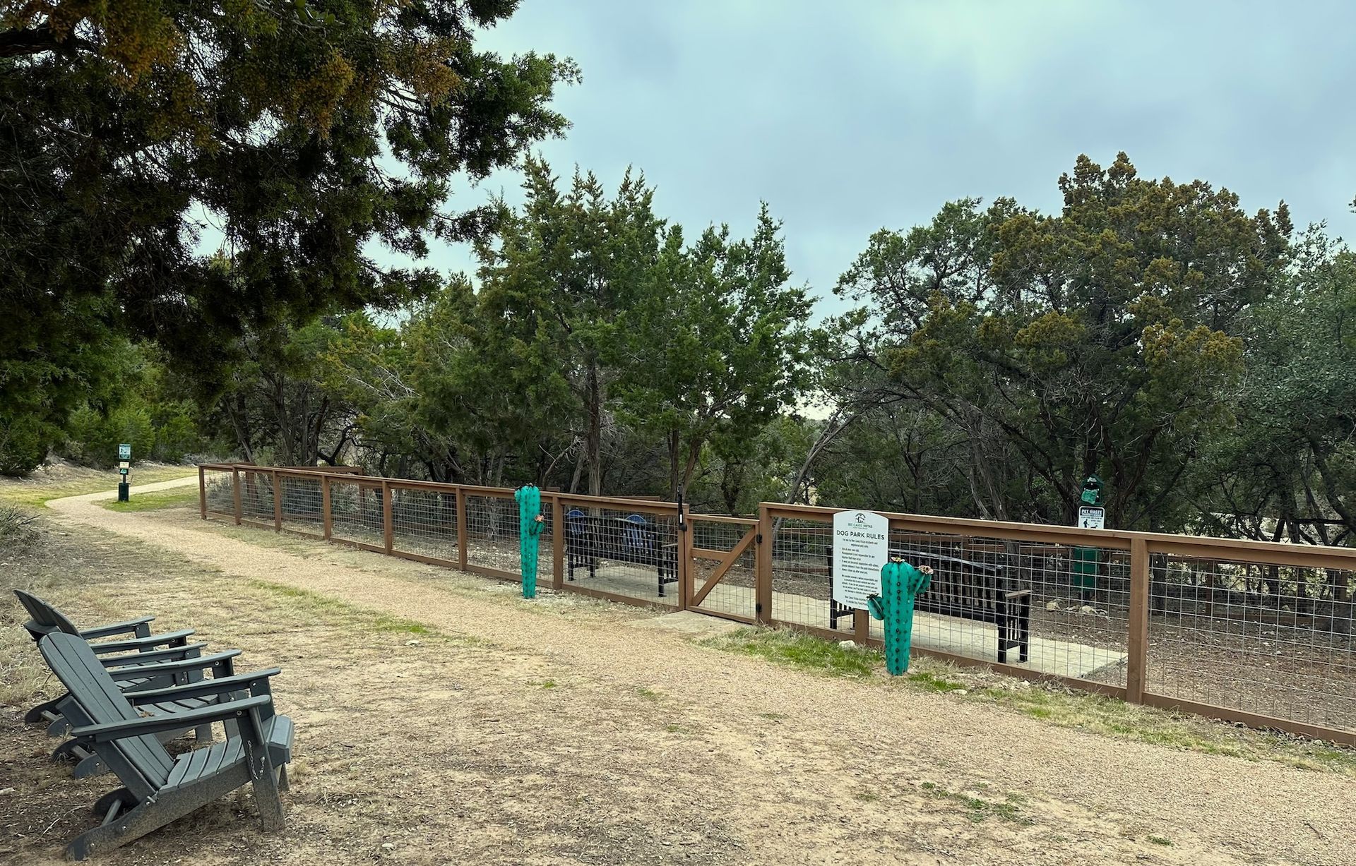Dog park with a gravel path, wooden fence, benches, and trees. Overcast sky.