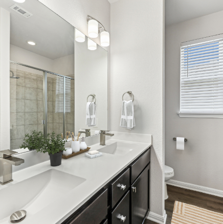 Bathroom with double sink vanity, large mirror, and shower. White countertops and walls, dark cabinets.