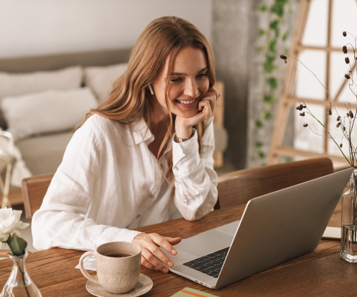Woman smiles while using laptop at a wooden table, coffee cup present.