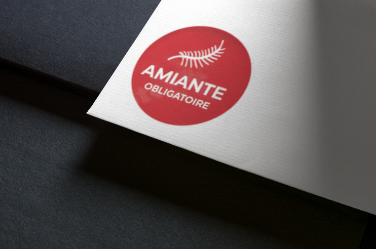 Cercle rouge avec texte blanc : « AMIANTÉ OBLIGATOIRE », avec un motif ressemblant à une fougère sur papier blanc sur fond noir.