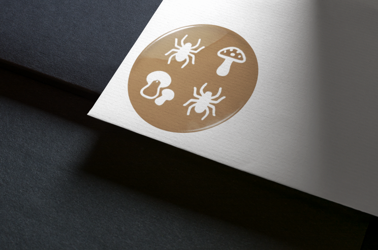 Autocollant rond doré avec des araignées et des champignons blancs sur une carte blanche, le tout sur une surface gris foncé.