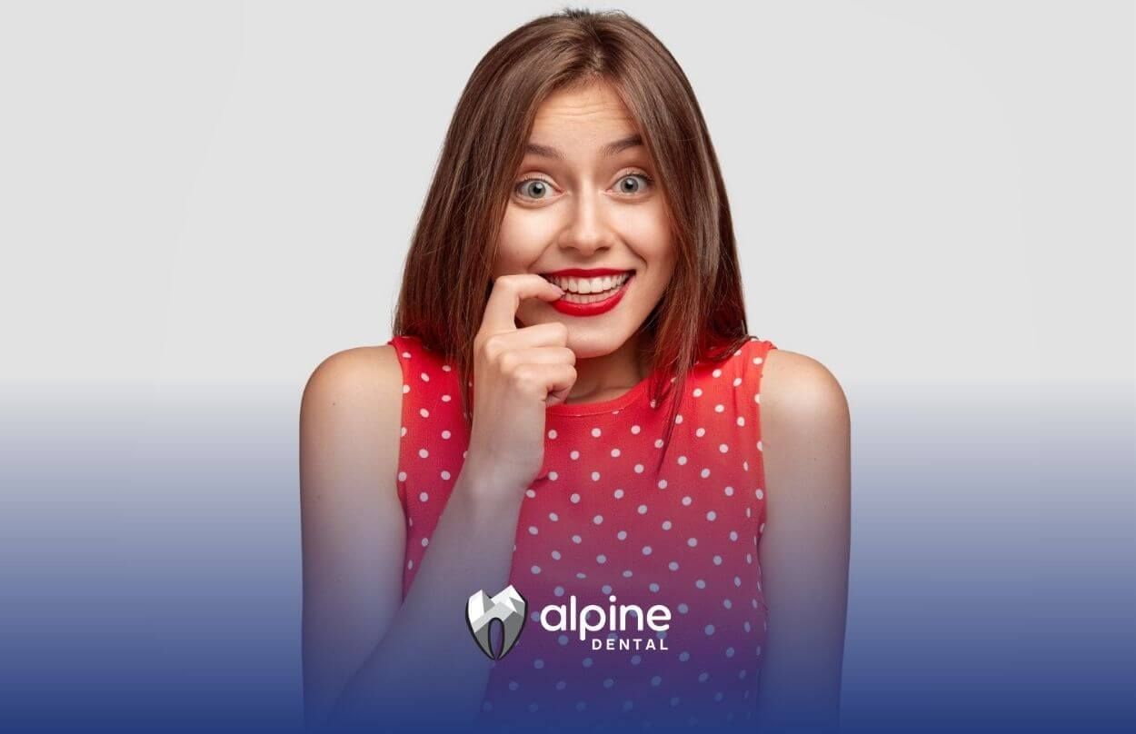Woman in red polka dot dress smiling nervously, biting her finger.
