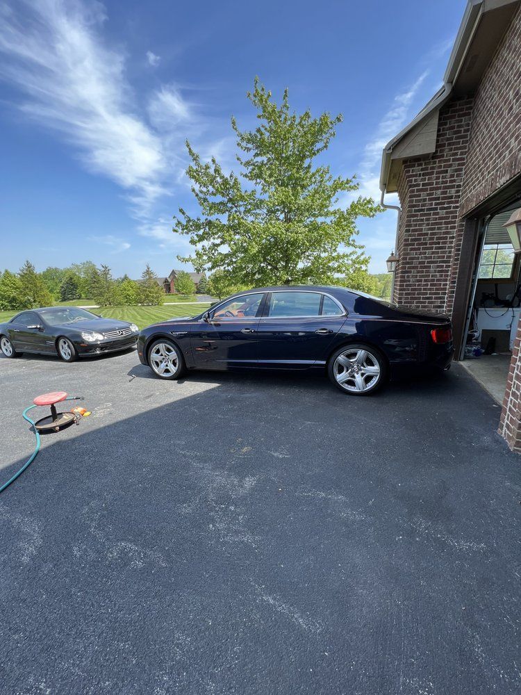 Dark blue Bentley parked next to a brick garage. Another car and a tree are visible. Sunny day.