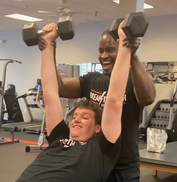 A man in a black shirt with the word mean on it is helping another man lift dumbbells