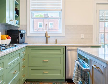 A kitchen with green cabinets , white counter tops , a sink , and a window.