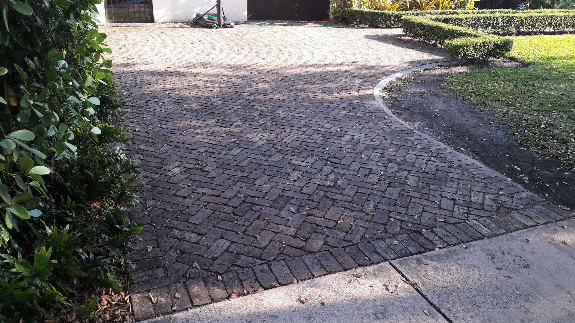 Brick driveway leading to a garage, bordered by a hedge and grass, with a sidewalk in the foreground.