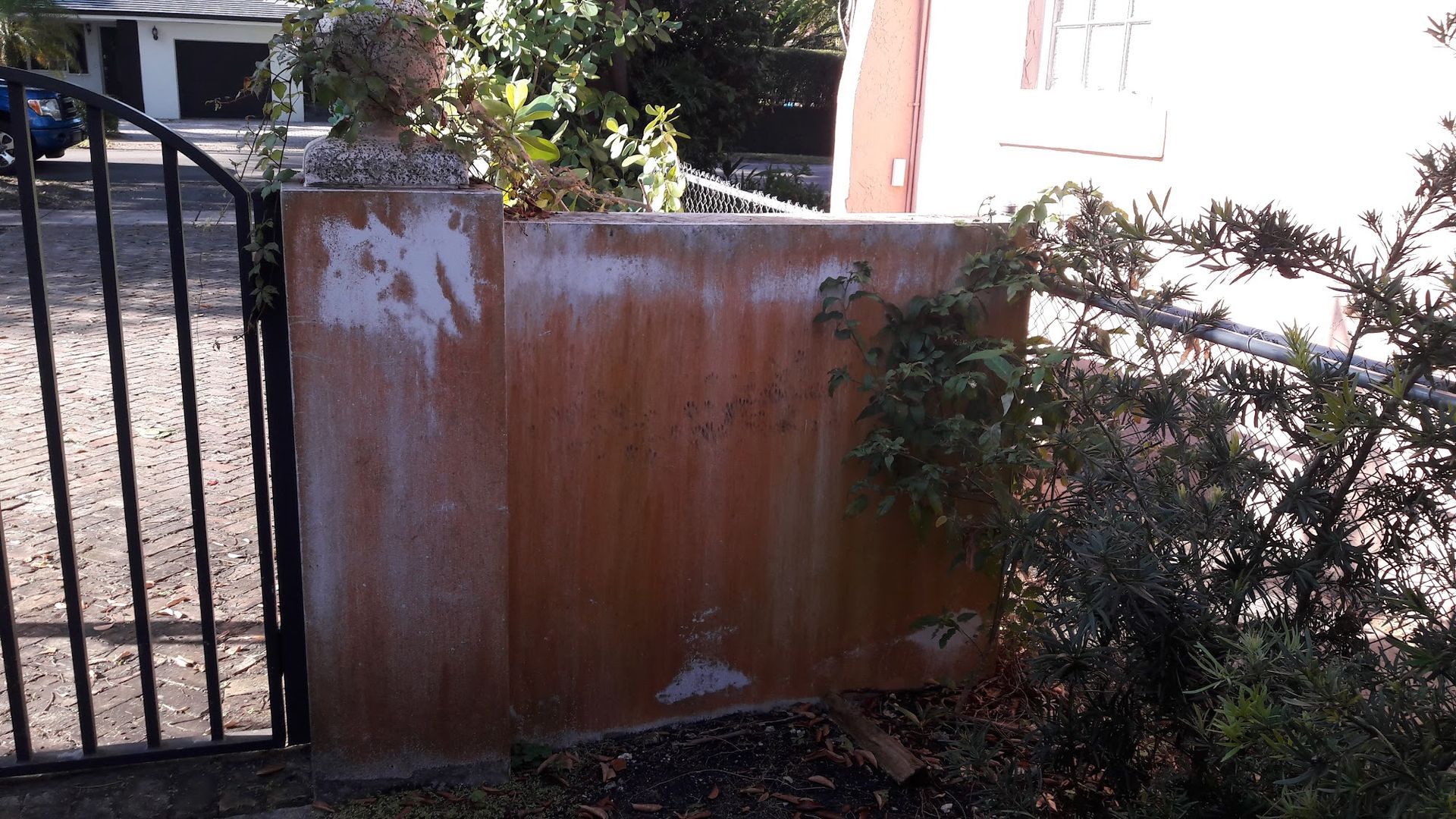 Brick wall with black gate and a decorative finial. Foliage grows around the wall, with a house visible in the background.