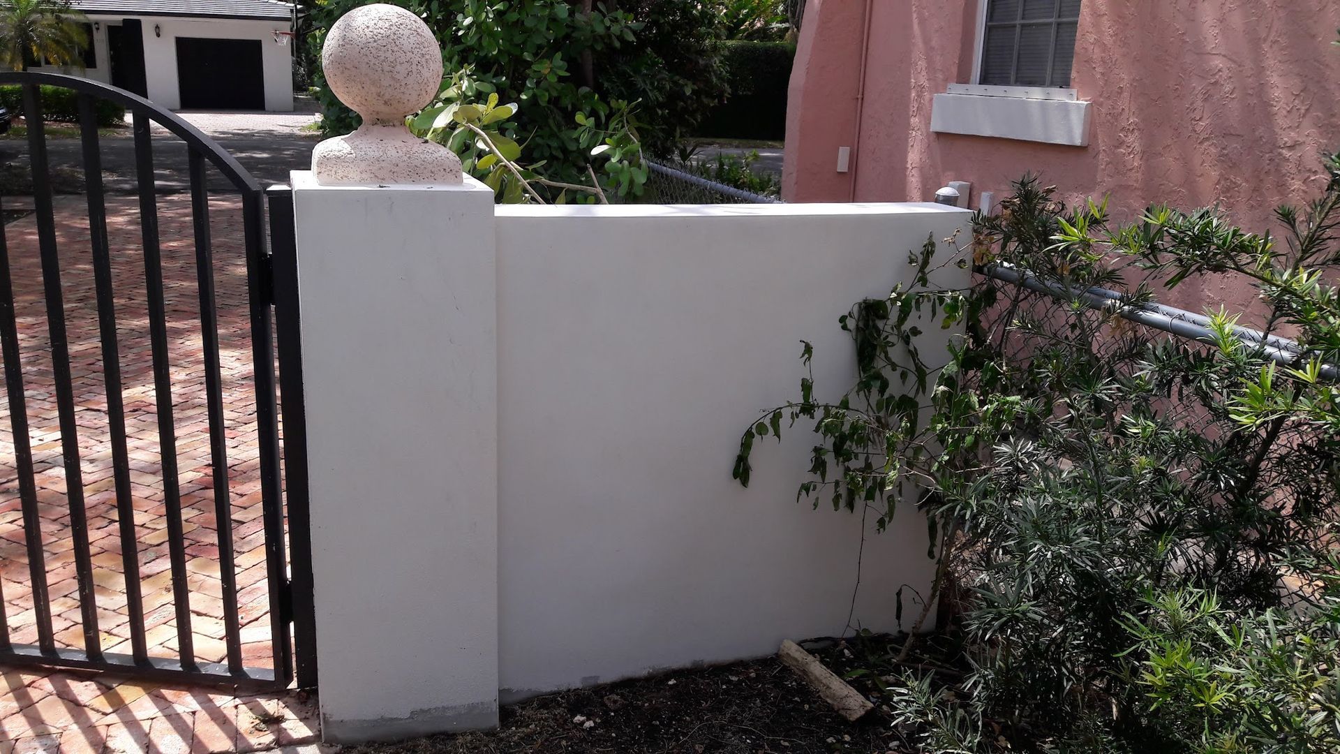 Black gate and wall with white pillars, a pink building in the background and lush greenery.