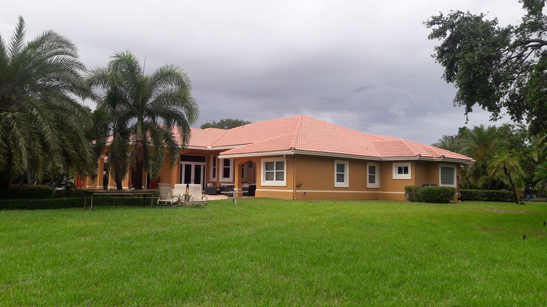 Single-story, tan house with a terracotta roof, set in a grassy yard with palm trees under a cloudy sky.
