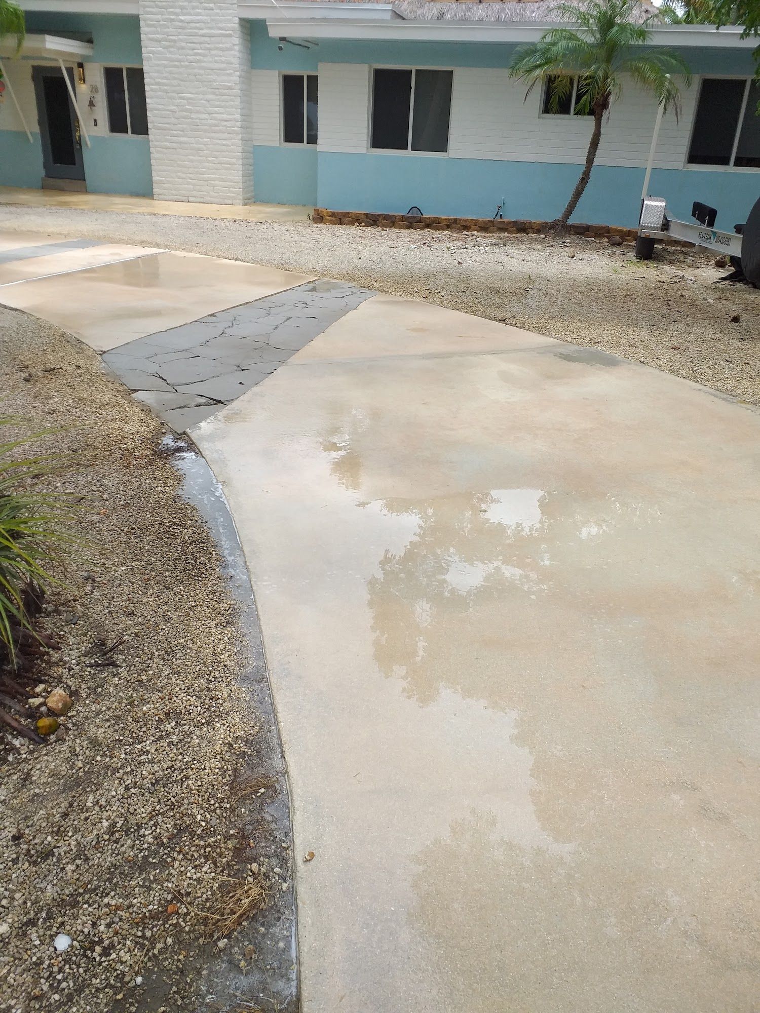 Wet concrete driveway leading to a light blue and white house, with gravel and landscaping on either side.
