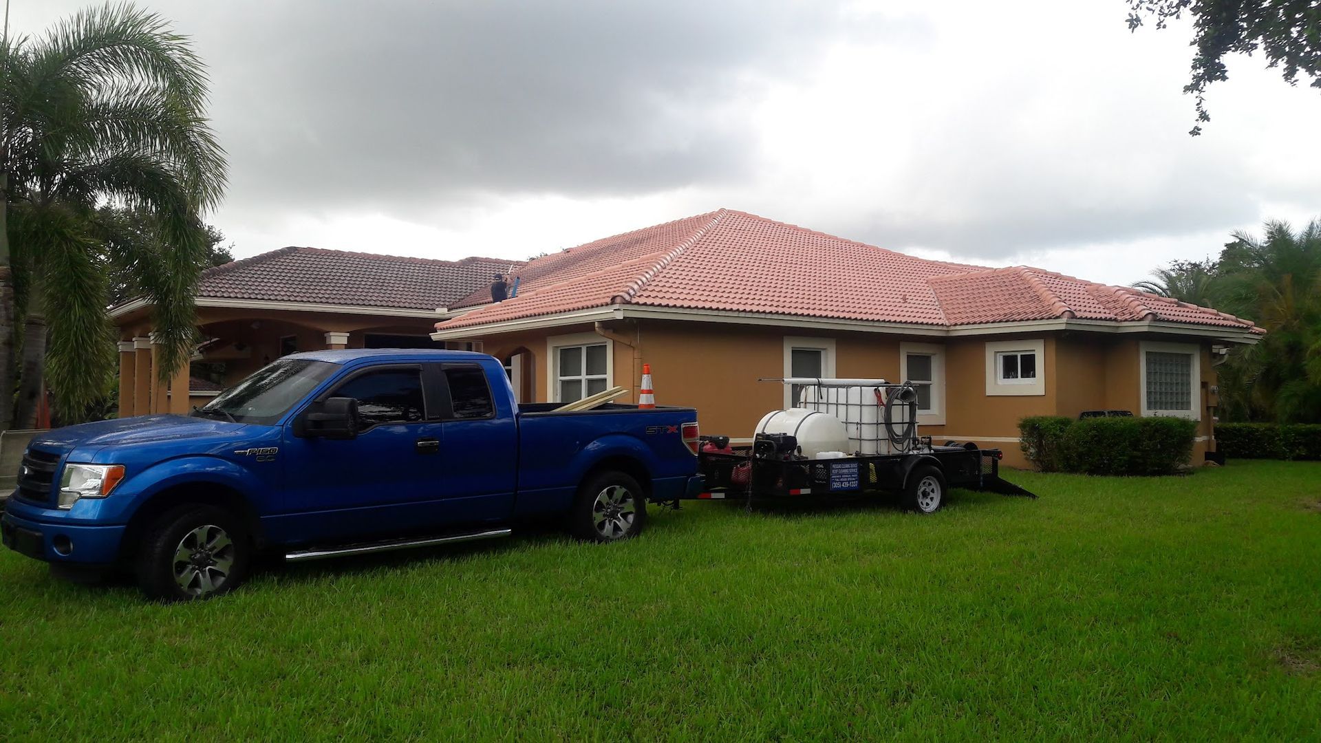Blue truck towing a trailer with cleaning equipment parked on a green lawn in front of a house with a red tile roof.