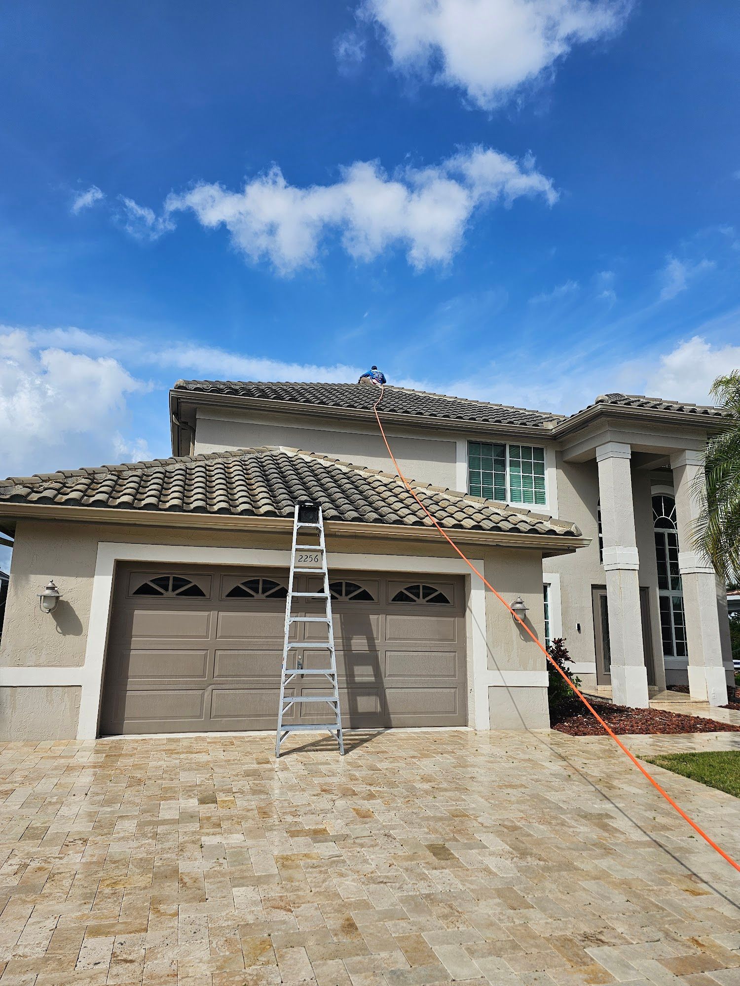 House with tan facade, brown garage doors, ladder on roof, blue sky.