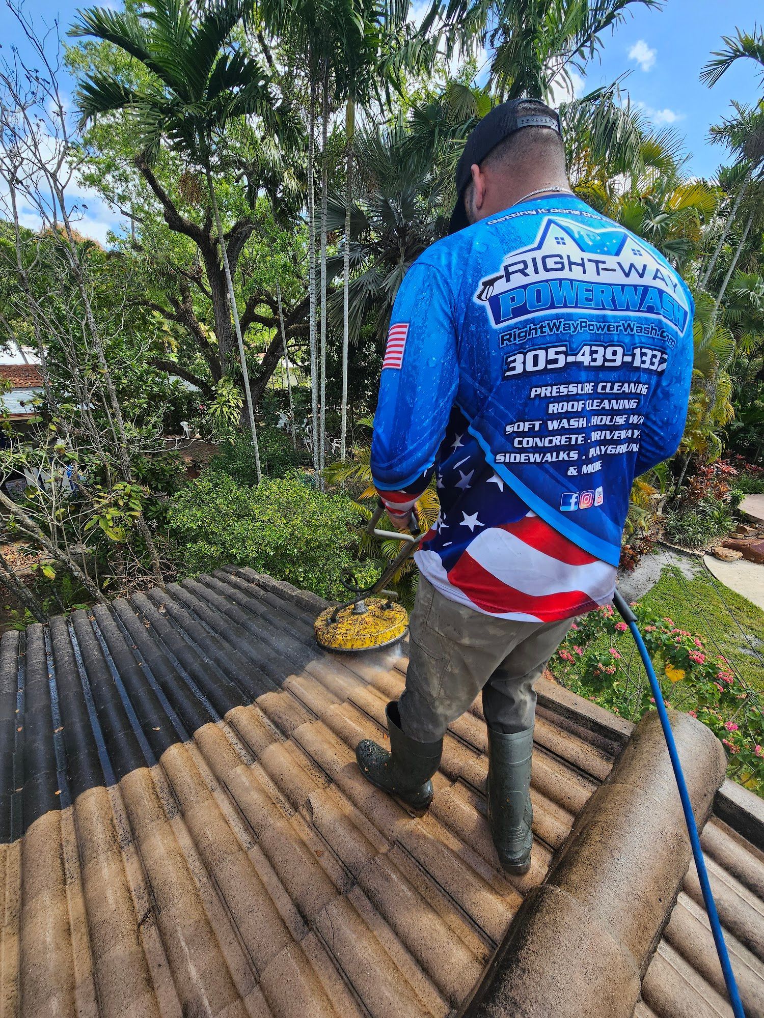 Man pressure washing a tile roof, wearing a blue shirt with company logo, outdoors.