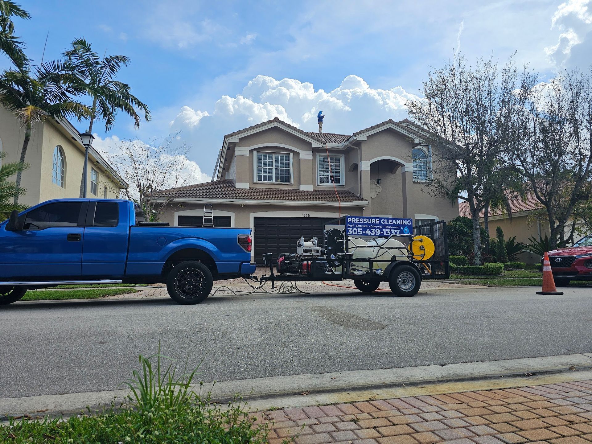 Blue truck towing a trailer parked in front of a house; workers are present, possibly cleaning the roof.