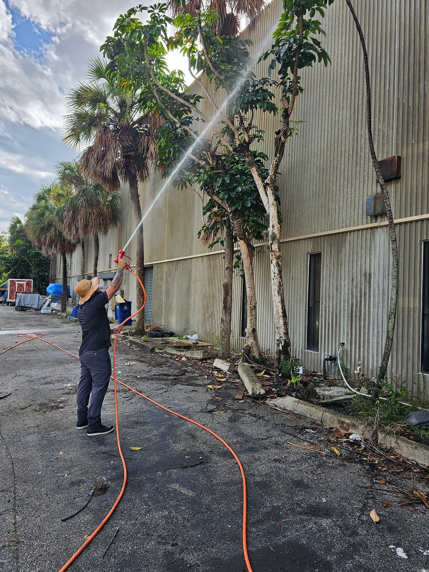 A person sprays water at a building exterior with a hose. Trees and debris are nearby.
