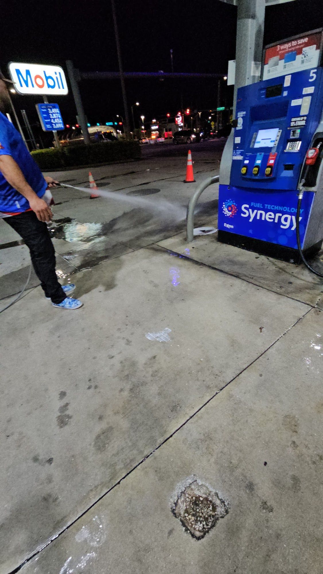 Person spraying water on a gas station concrete floor near a pump at night.