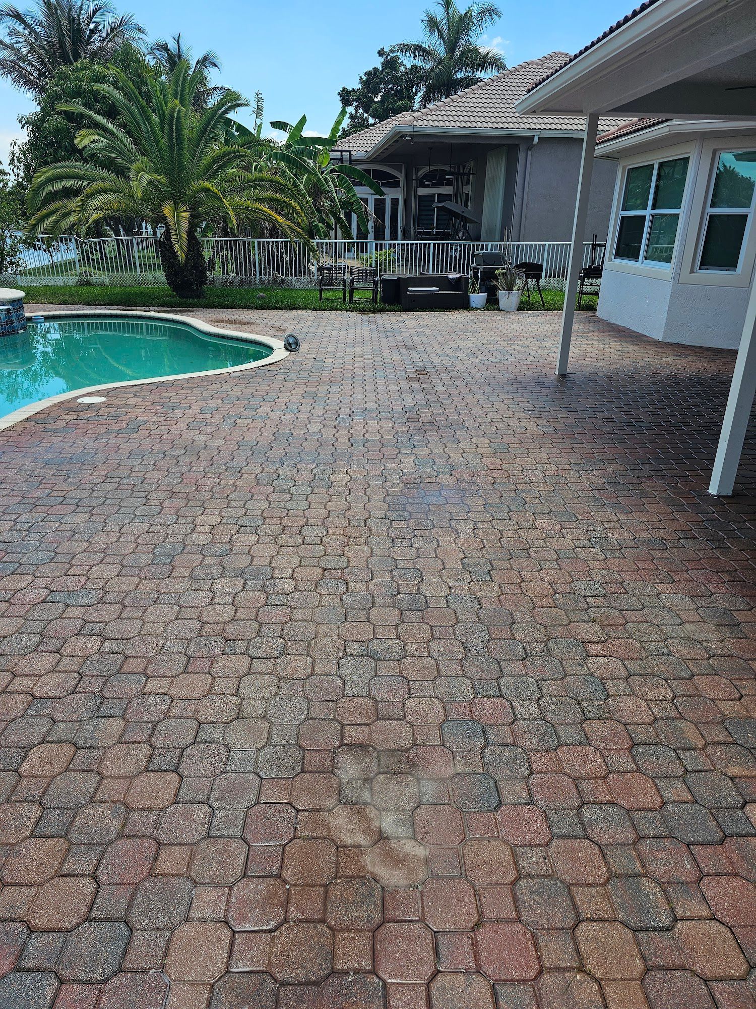 Brick patio beside a pool and house, with palm trees under a blue sky.
