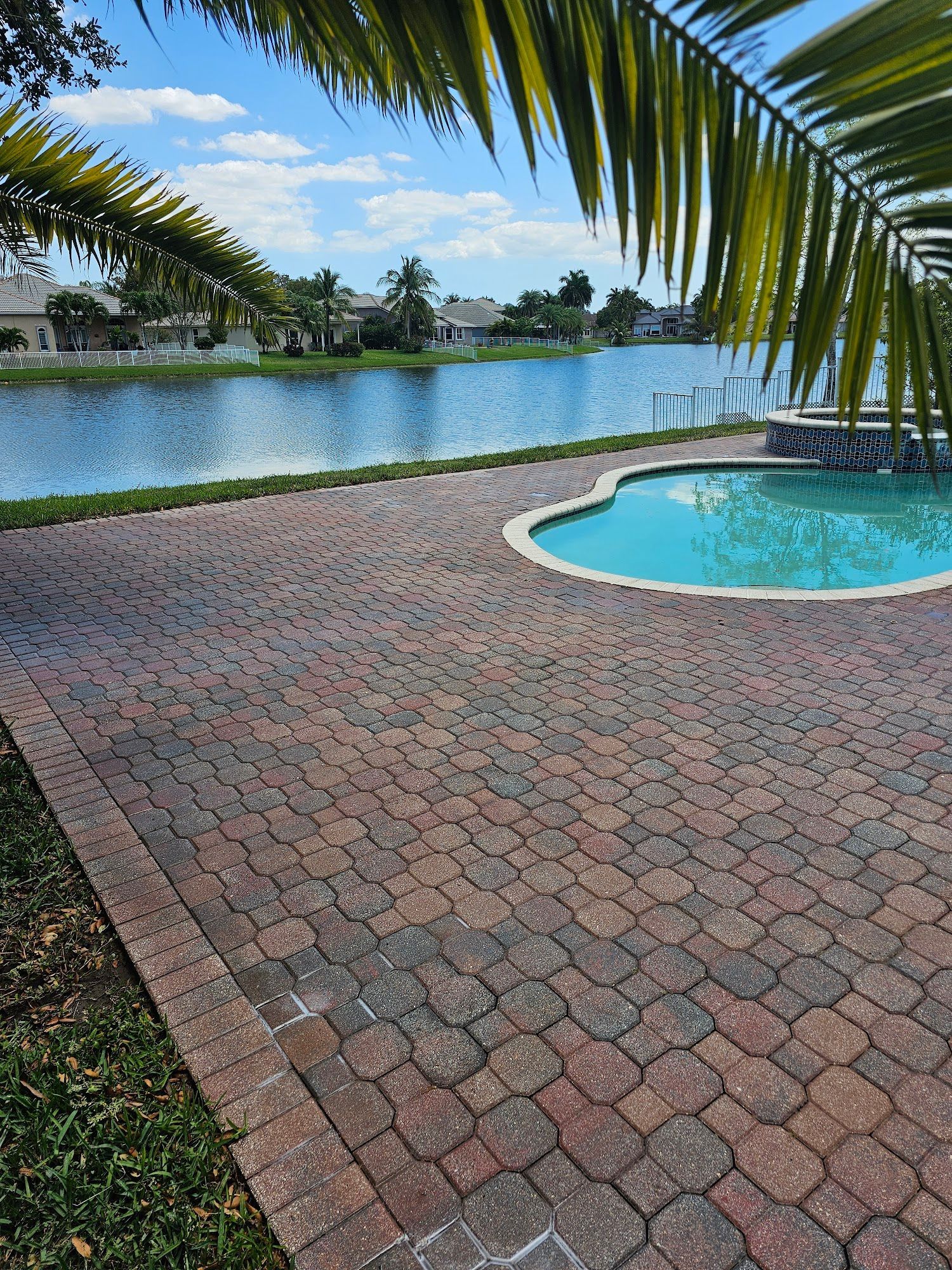 Patio overlooking a lake and pool, framed by palm leaves. Brick pavers and blue water.