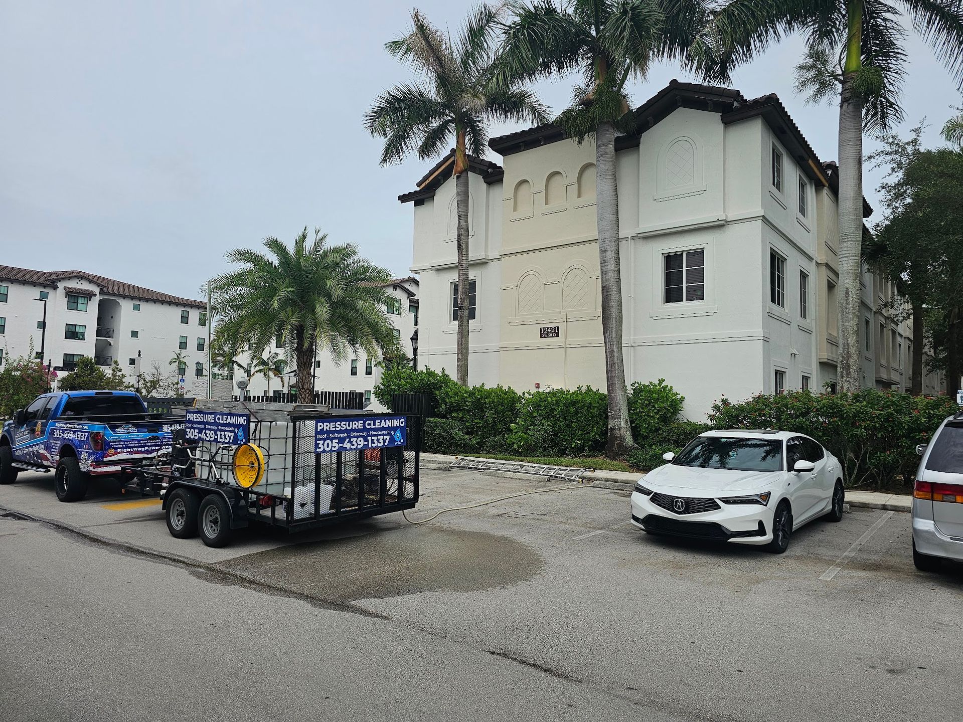 A power washing truck and trailer parked near a white apartment building and cars.