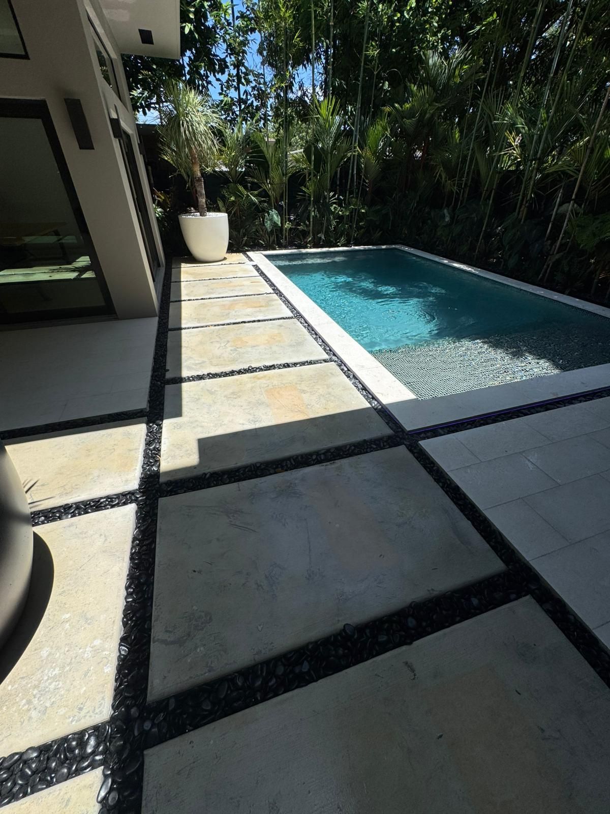 Poolside patio with a small rectangular pool, surrounded by square tiles and gravel. Lush greenery in the background.