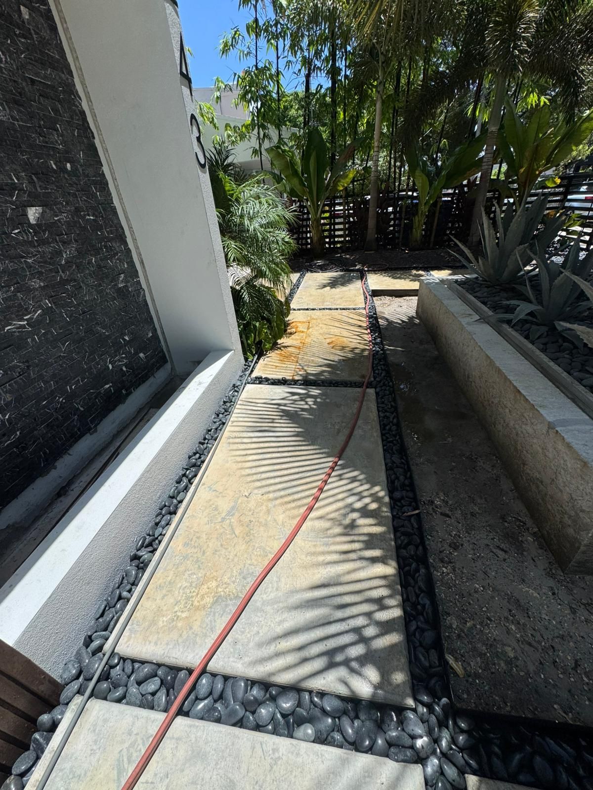 Concrete path flanked by black stones and a narrow garden bed, leading into a sunny backyard.