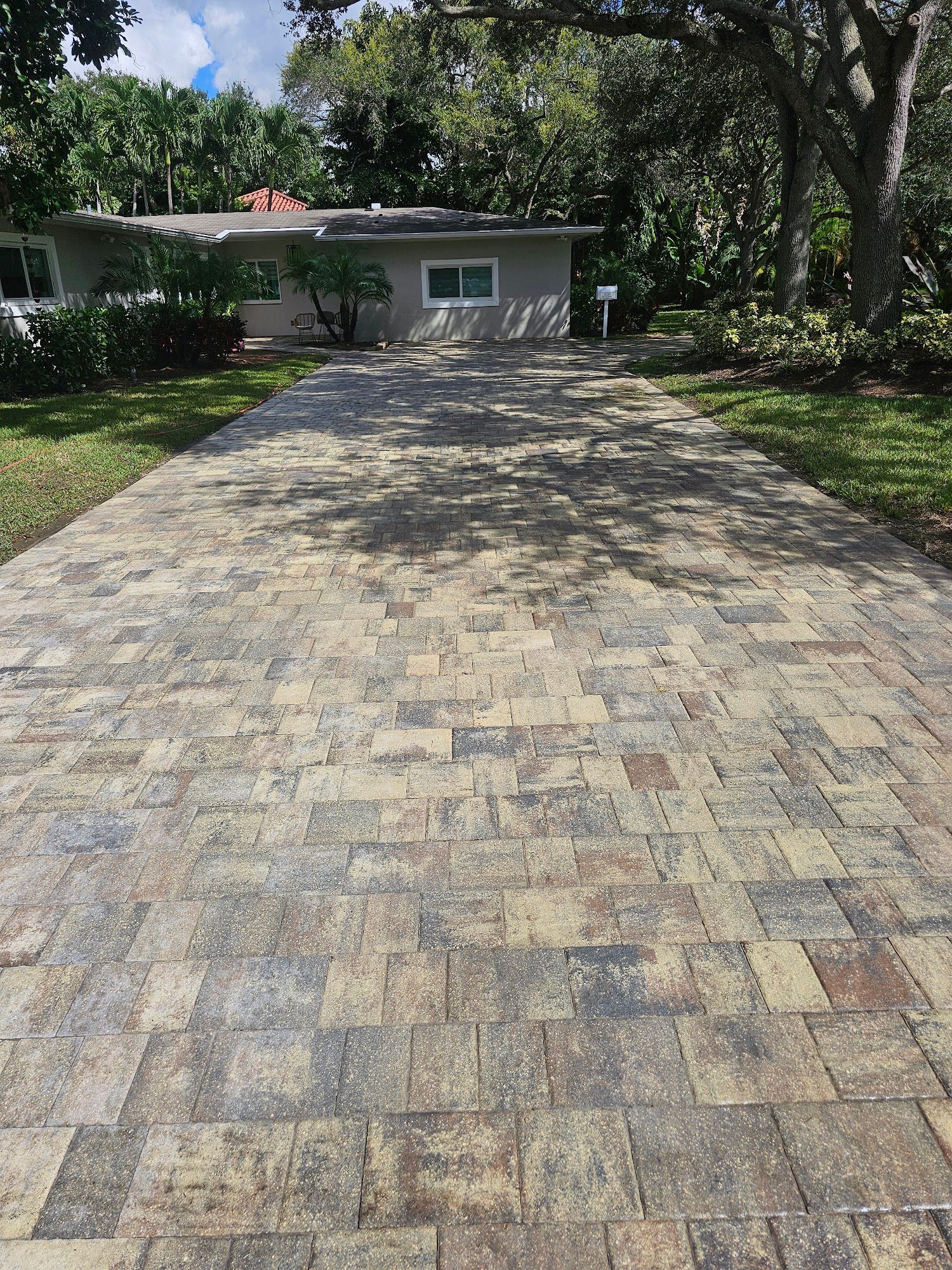 Paver driveway leading to a gray house with landscaping; sunny day.